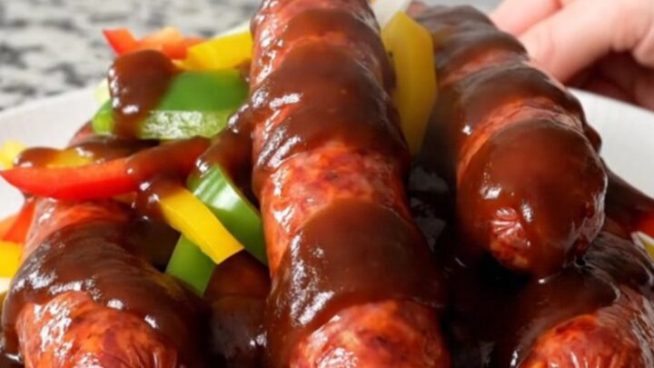 A close-up of hot link sausages in a slow cooker topped with colorful bell peppers and onions in barbecue sauce.