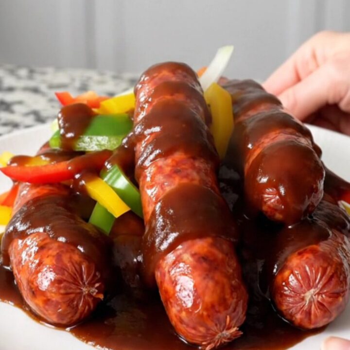 A close-up of hot link sausages in a slow cooker topped with colorful bell peppers and onions in barbecue sauce.