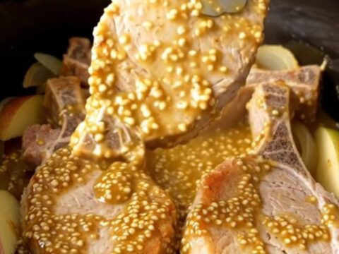 A close-up of tender, cooked pork chops topped with a golden mustard sauce, onions, and apple wedges in a slow cooker.