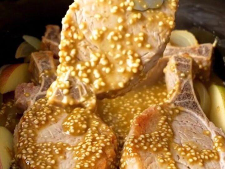 A close-up of tender, cooked pork chops topped with a golden mustard sauce, onions, and apple wedges in a slow cooker.