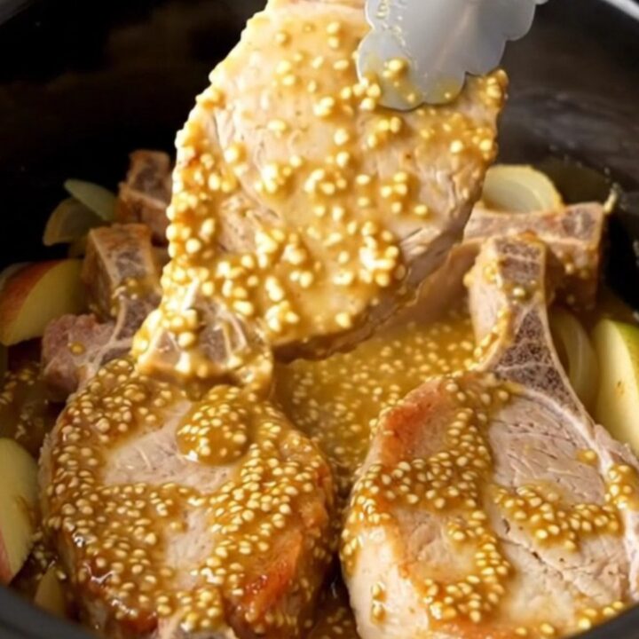 A close-up of tender, cooked pork chops topped with a golden mustard sauce, onions, and apple wedges in a slow cooker.