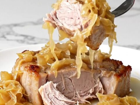 A close-up of tender, slow-cooked pork steaks topped with sauerkraut and sliced apples on a white plate.