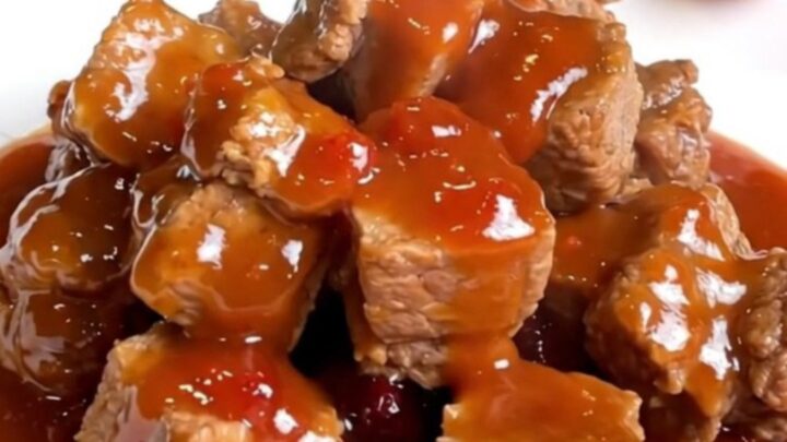 A close-up of tender, bite-sized pork loin cubes coated in a thick, glossy, reddish-purple grape jelly and chili glaze.