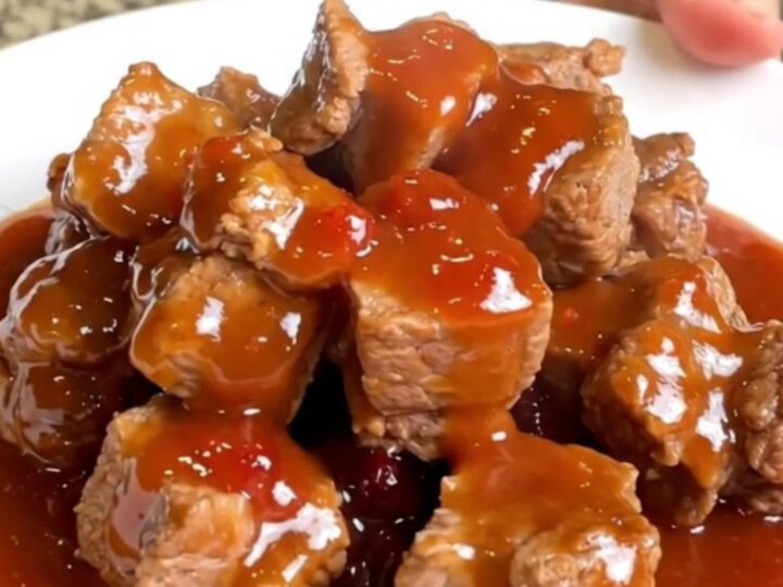 A close-up of tender, bite-sized pork loin cubes coated in a thick, glossy, reddish-purple grape jelly and chili glaze.