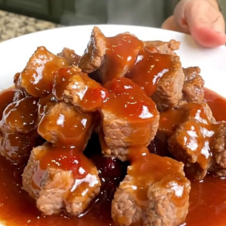 A close-up of tender, bite-sized pork loin cubes coated in a thick, glossy, reddish-purple grape jelly and chili glaze.