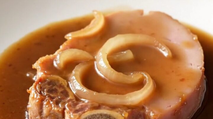 A close-up of a tender bone-in ham steak glazed in a dark, rich coffee gravy with soft onions on a white plate.