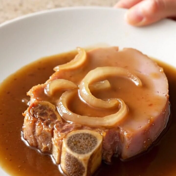 A close-up of a tender bone-in ham steak glazed in a dark, rich coffee gravy with soft onions on a white plate.