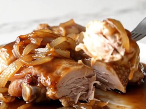 A close-up of slow-cooked chicken thighs in a dark, rich sauce with onions, served on a white plate.