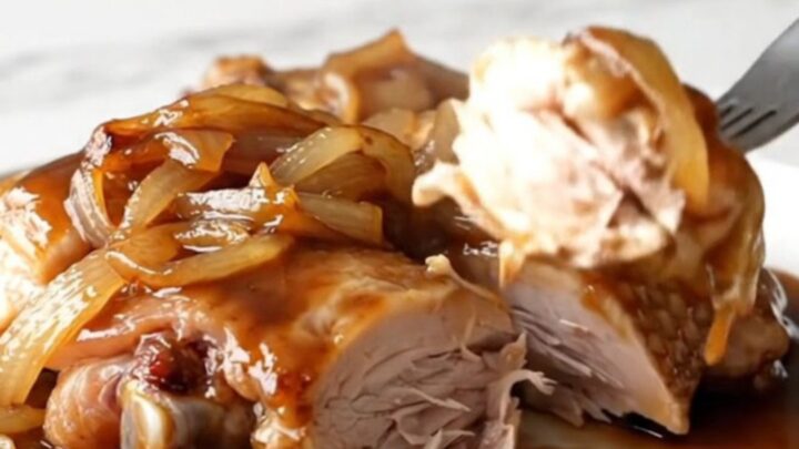 A close-up of slow-cooked chicken thighs in a dark, rich sauce with onions, served on a white plate.