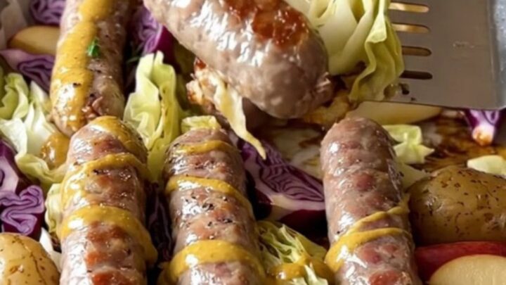 A sheet pan filled with golden-brown bratwurst, purple cabbage wedges, halved baby potatoes, and roasted apples.