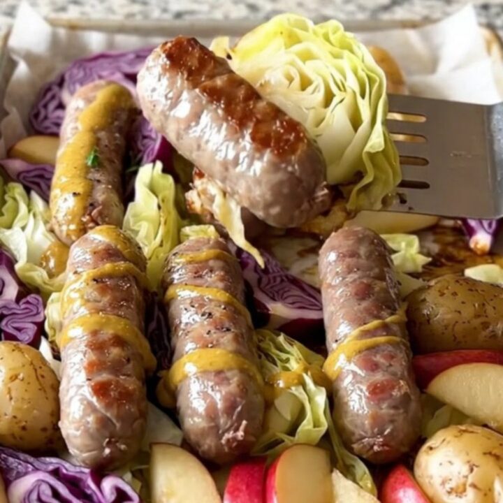 A sheet pan filled with golden-brown bratwurst, purple cabbage wedges, halved baby potatoes, and roasted apples.