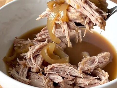 Tender shredded pork shoulder in a dark, savory au jus with soft onions in a slow cooker.