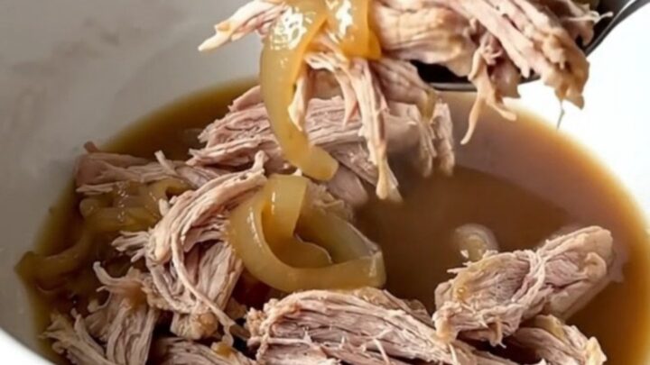 Tender shredded pork shoulder in a dark, savory au jus with soft onions in a slow cooker.