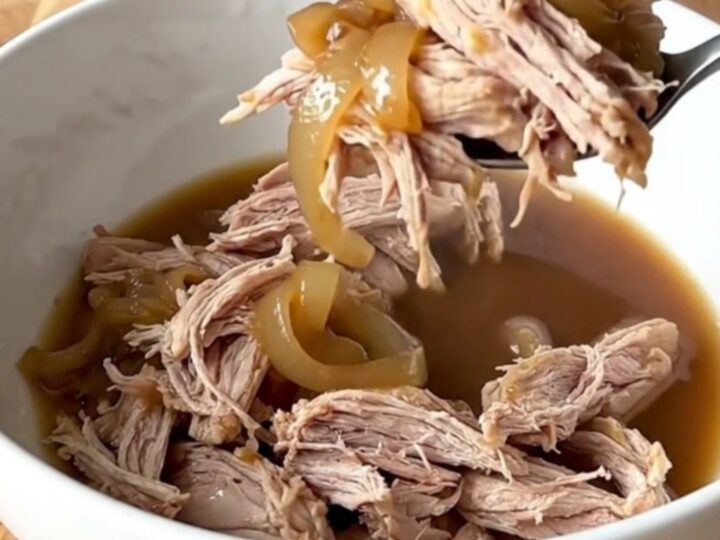 Tender shredded pork shoulder in a dark, savory au jus with soft onions in a slow cooker.
