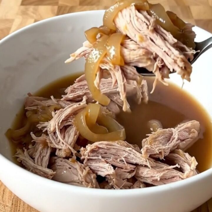 Tender shredded pork shoulder in a dark, savory au jus with soft onions in a slow cooker.