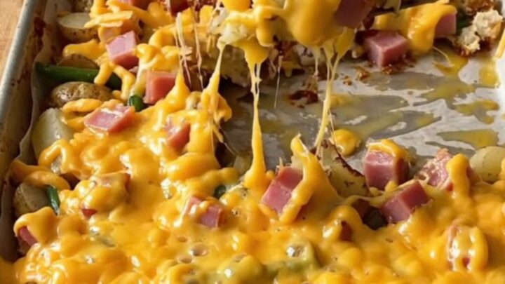 A golden sheet pan filled with roasted ham cubes, baby potatoes, and green beans topped with melted cheddar cheese.