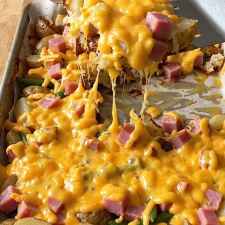 A golden sheet pan filled with roasted ham cubes, baby potatoes, and green beans topped with melted cheddar cheese.