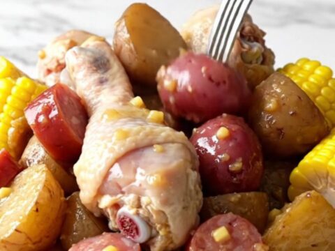 A close-up of a slow cooker filled with chicken drumsticks, corn, potatoes, and sausage in a golden butter sauce.