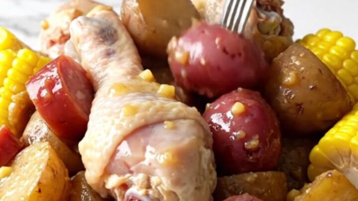 A close-up of a slow cooker filled with chicken drumsticks, corn, potatoes, and sausage in a golden butter sauce.
