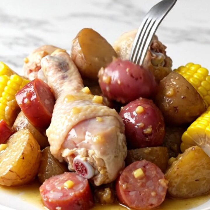 A close-up of a slow cooker filled with chicken drumsticks, corn, potatoes, and sausage in a golden butter sauce.