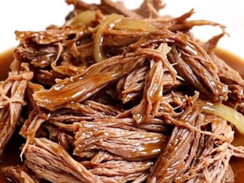 A close-up of a slow cooker filled with shredded beef chuck roast in a rich, dark gravy with onions and garlic.
