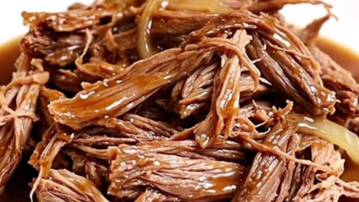 A close-up of a slow cooker filled with shredded beef chuck roast in a rich, dark gravy with onions and garlic.