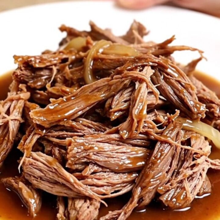 A close-up of a slow cooker filled with shredded beef chuck roast in a rich, dark gravy with onions and garlic.
