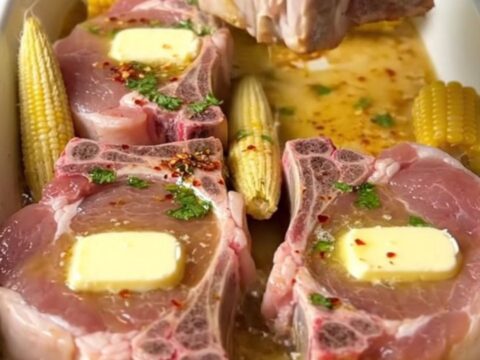 A large ceramic baking dish filled with golden-brown bone-in pork chops and mini corn cobs in a cilantro lime butter sauce.