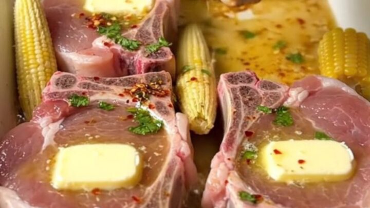 A large ceramic baking dish filled with golden-brown bone-in pork chops and mini corn cobs in a cilantro lime butter sauce.