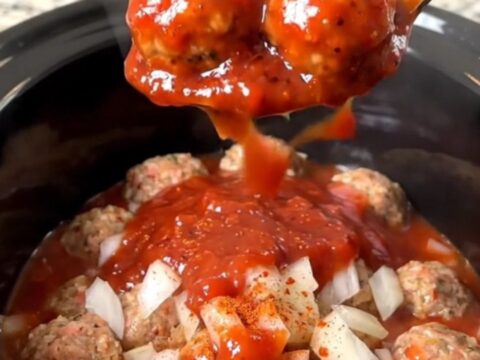 A close-up view of slow cooker strawberry BBQ meatballs coated in a glossy, red sauce in a black ceramic pot.