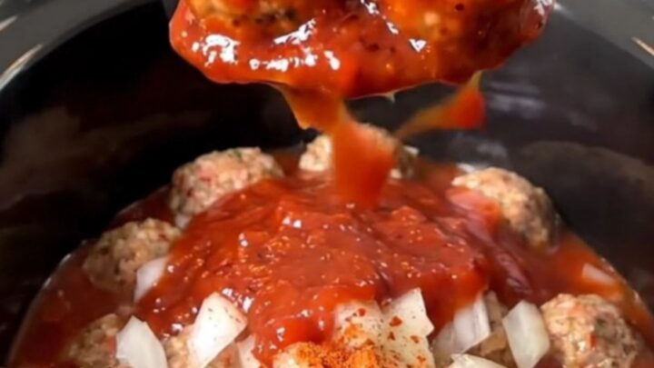 A close-up view of slow cooker strawberry BBQ meatballs coated in a glossy, red sauce in a black ceramic pot.