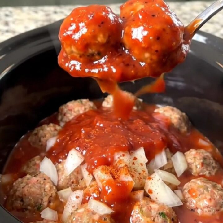 A close-up view of slow cooker strawberry BBQ meatballs coated in a glossy, red sauce in a black ceramic pot.