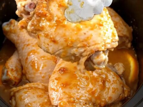 A close-up of tender, braised chicken thighs in a slow cooker, garnished with fresh orange slices and aromatic rosemary.