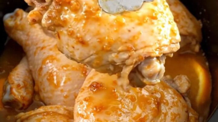 A close-up of tender, braised chicken thighs in a slow cooker, garnished with fresh orange slices and aromatic rosemary.