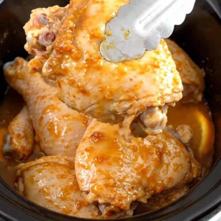 A close-up of tender, braised chicken thighs in a slow cooker, garnished with fresh orange slices and aromatic rosemary.