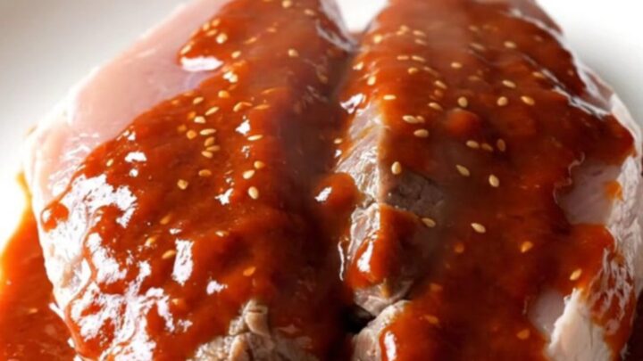A slow-cooked pork tenderloin with a glossy, deep red gochujang honey glaze on a white plate.