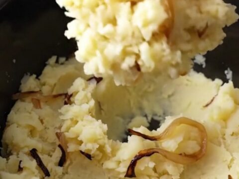 Slow cooker filled with chunky, rustic mashed potatoes with visible tender onion strands in a bright kitchen.