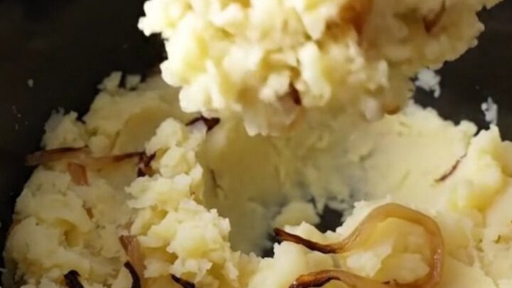 Slow cooker filled with chunky, rustic mashed potatoes with visible tender onion strands in a bright kitchen.