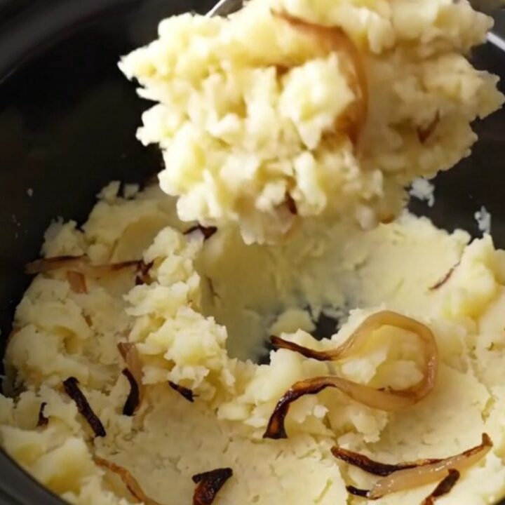 Slow cooker filled with chunky, rustic mashed potatoes with visible tender onion strands in a bright kitchen.