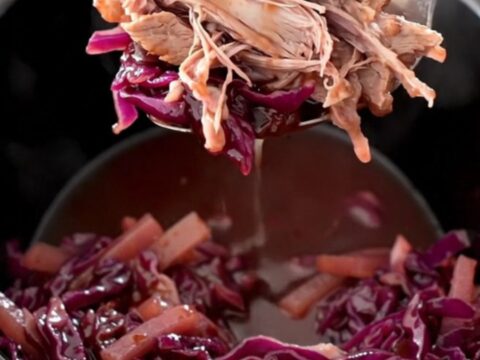 A rustic slow cooker filled with tender chunks of pork shoulder, purple red cabbage, and sliced apples in a glossy sauce.