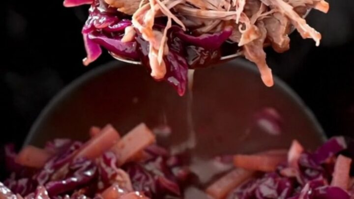 A rustic slow cooker filled with tender chunks of pork shoulder, purple red cabbage, and sliced apples in a glossy sauce.
