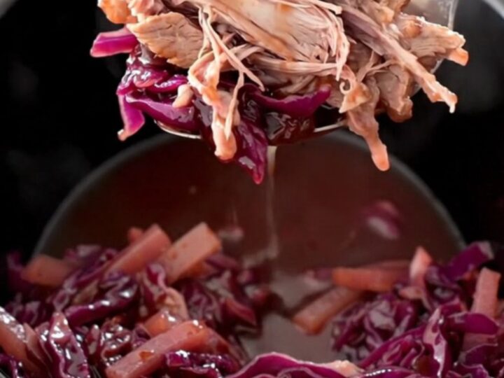 A rustic slow cooker filled with tender chunks of pork shoulder, purple red cabbage, and sliced apples in a glossy sauce.