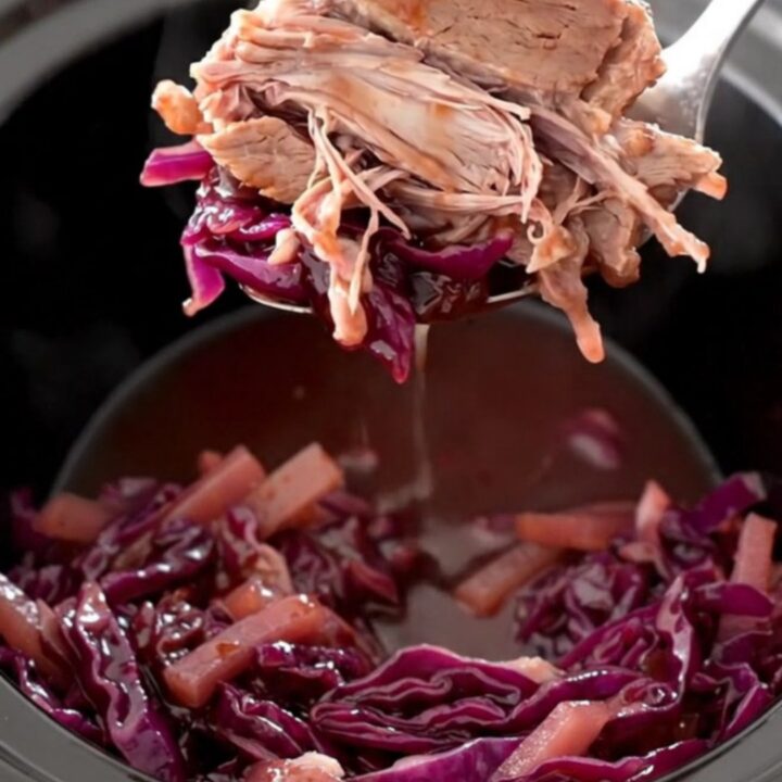 A rustic slow cooker filled with tender chunks of pork shoulder, purple red cabbage, and sliced apples in a glossy sauce.