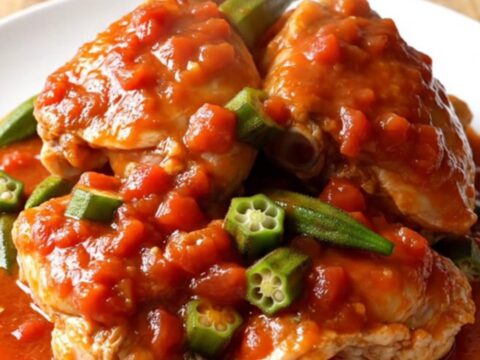 A close-up of tender chicken thighs and sliced okra in a rich, thick red tomato sauce inside a white slow cooker.