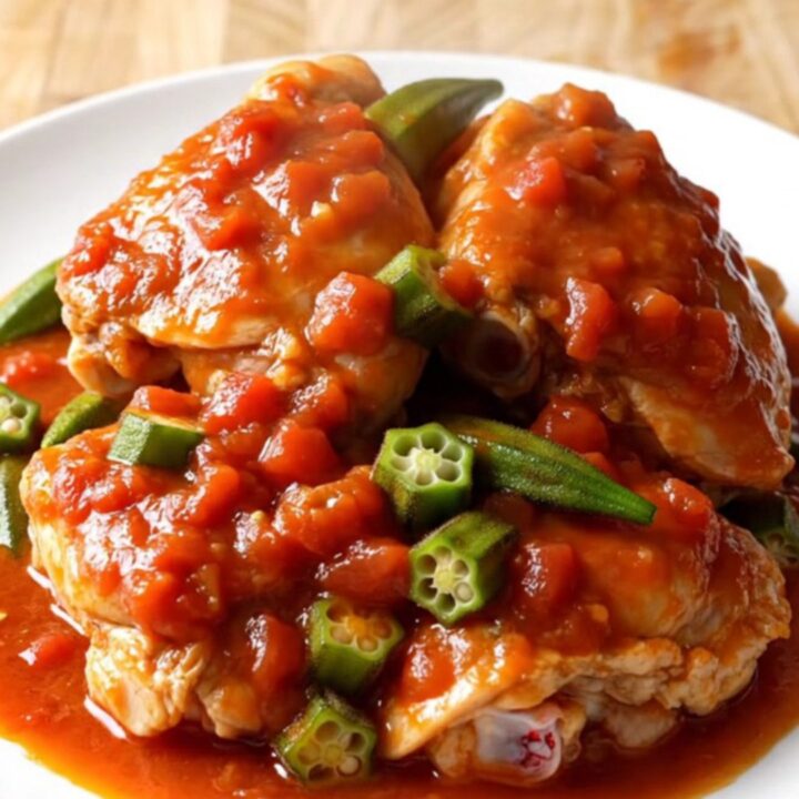 A close-up of tender chicken thighs and sliced okra in a rich, thick red tomato sauce inside a white slow cooker.