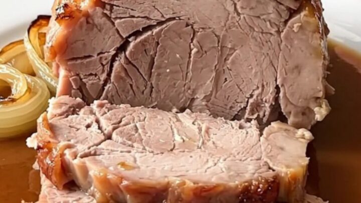 A whole, cooked boneless pork loin roast sitting in a white ceramic slow cooker insert with sliced yellow onions.