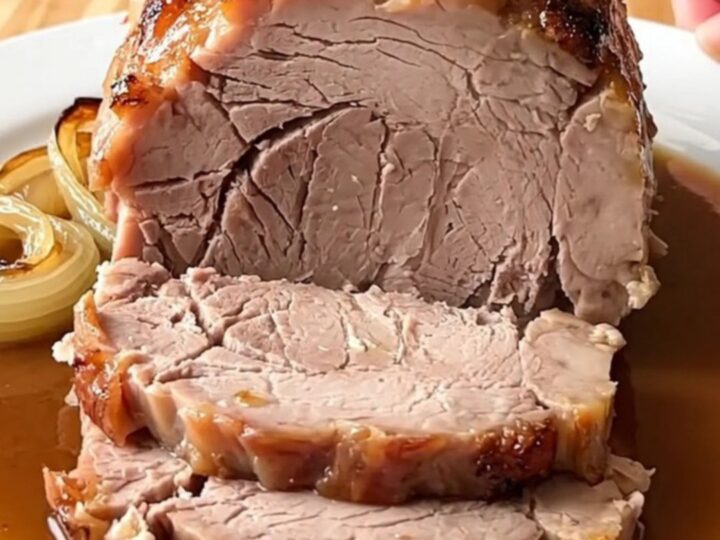 A whole, cooked boneless pork loin roast sitting in a white ceramic slow cooker insert with sliced yellow onions.