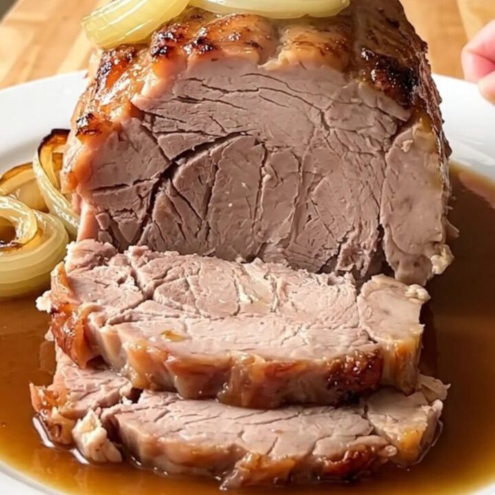 A whole, cooked boneless pork loin roast sitting in a white ceramic slow cooker insert with sliced yellow onions.