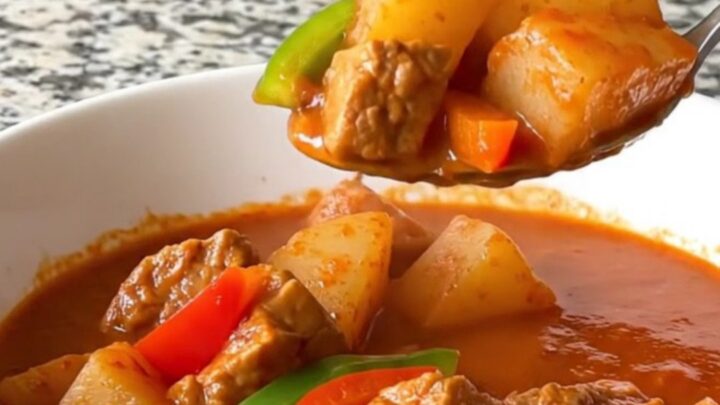A close-up of a slow cooker filled with cubes of pork, potatoes, and peppers in a rich tomato sauce.