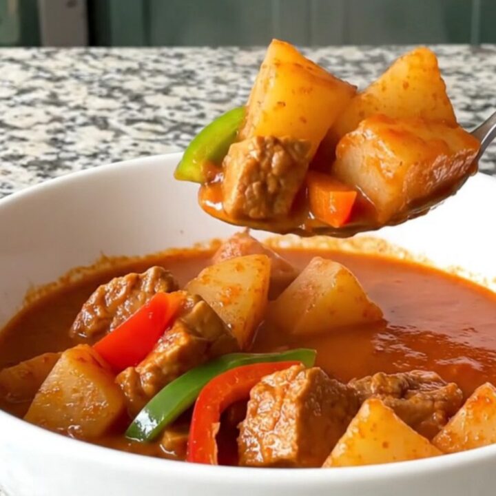 A close-up of a slow cooker filled with cubes of pork, potatoes, and peppers in a rich tomato sauce.